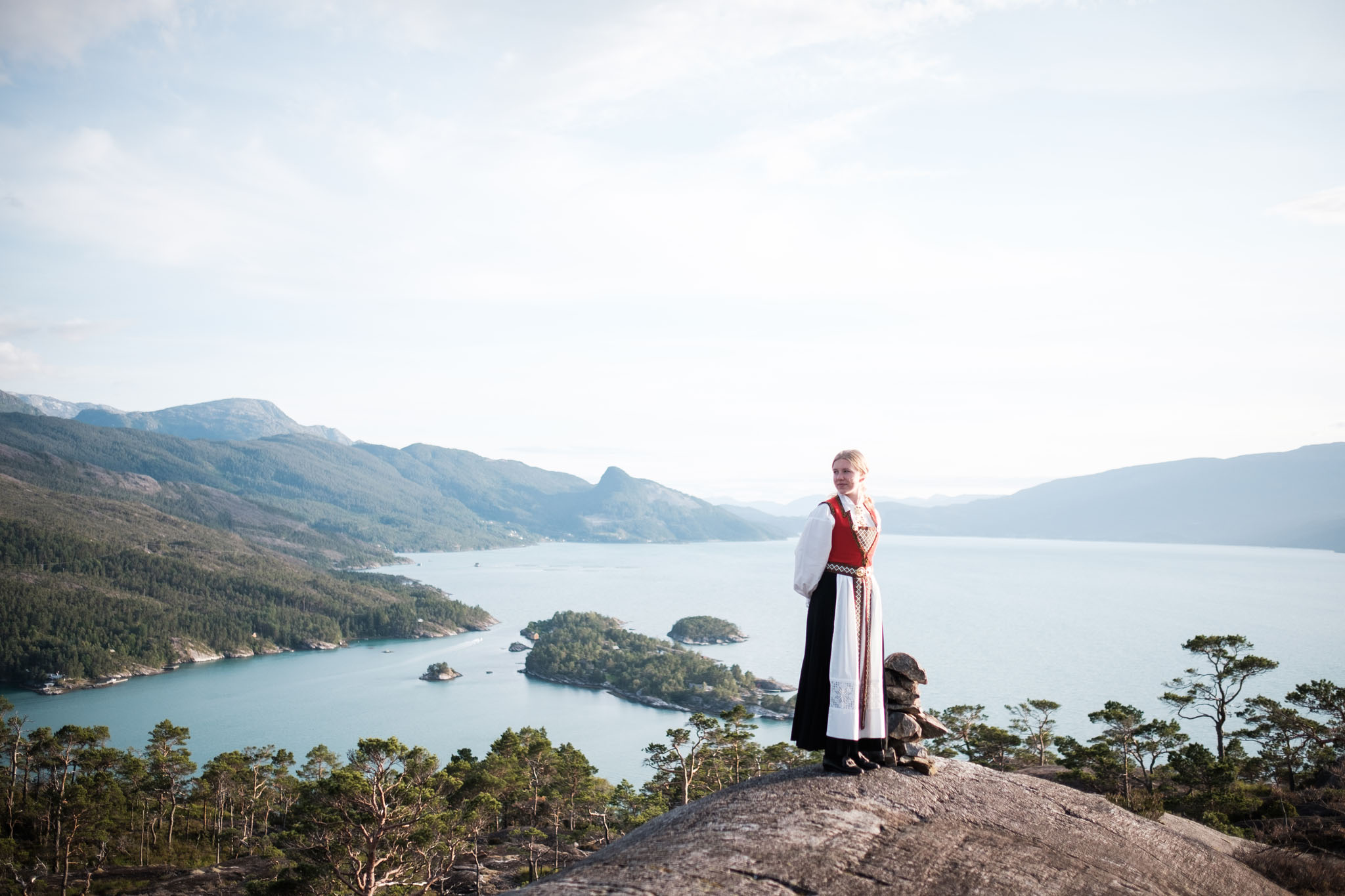 Konfirmant som står foran et flott fjordlandskap i Hardanger. Ikledd hardangerbunad.